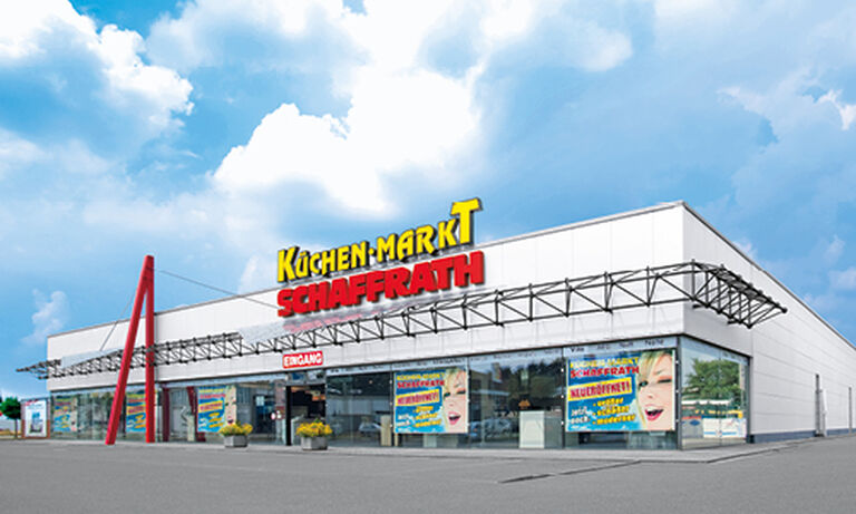 Das Bild zeigt den „Küchen-Markt Schaffrath“, ein modernes Gebäude mit einer weißen Fassade und einem klar strukturierten Design. Auf dem Dach befindet sich der Schriftzug „KÜCHEN-MARKT“ in gelben und weißen Buchstaben mit einem großen roten „SCHAFFRATH“ darunter. Der Eingangsbereich ist durch einen überdachten Vorbau mit einer Metallkonstruktion gekennzeichnet. Links vom Eingang befinden sich zwei große rote Säulen, die die Architektur optisch betonen. Die Glasfront des Gebäudes zeigt mehrere bunte Werbeplakate mit fröhlichen Gesichtern und Slogans, die den Küchenmarkt bewerben. Im Vordergrund sind ein gepflasterter Parkplatz sowie Pflanzkübel mit gelben Blumen zu sehen. Der Himmel ist hellblau mit weißen Wolken, was eine einladende und freundliche Atmosphäre schafft.